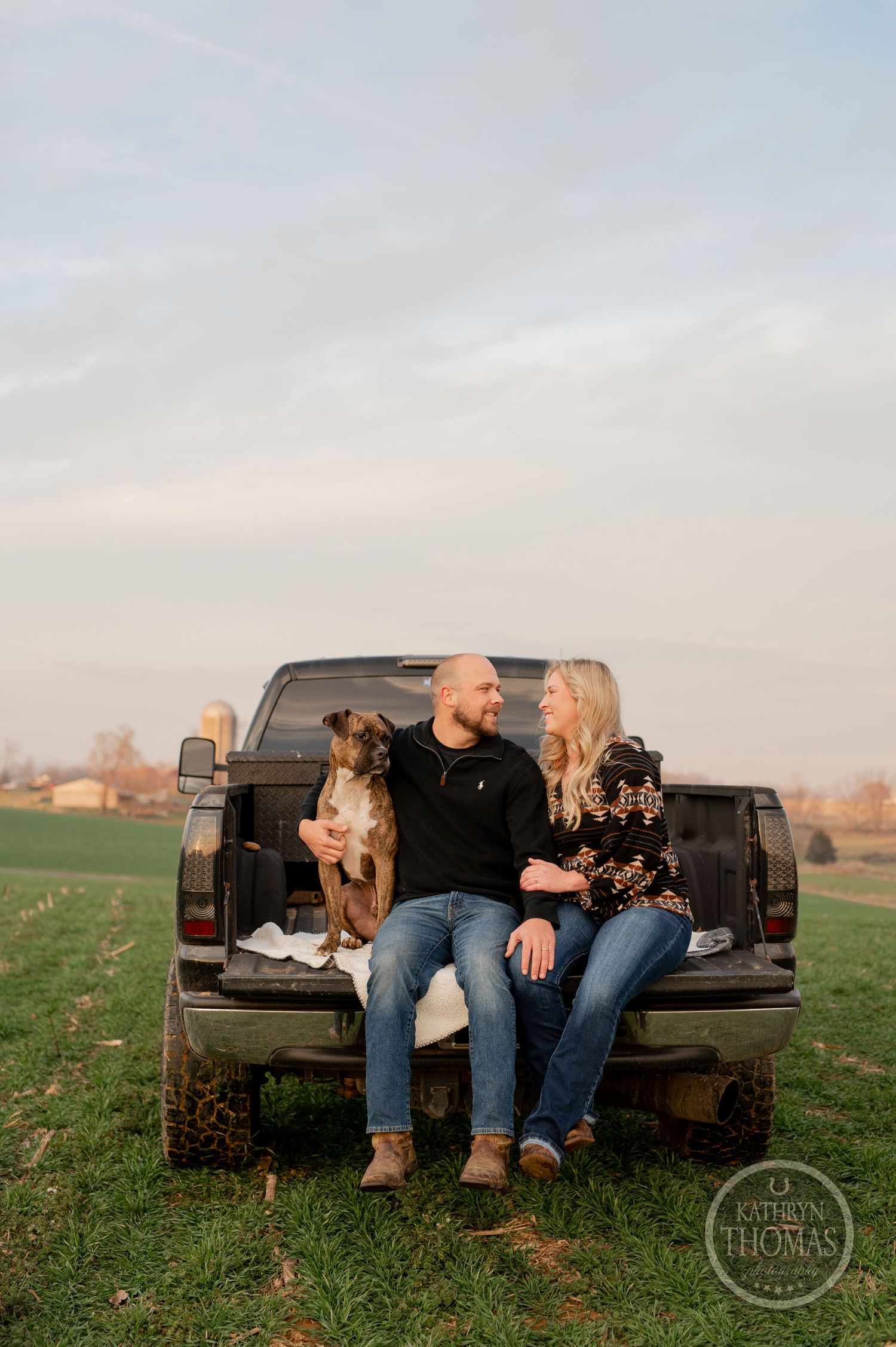 A couple sits on a pickup truck tailgate with their two dogs against a rural sunset backdrop.