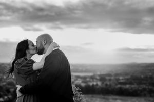 A black and white photo captures an intimate embrace against a dramatic cloudy sky and rolling hills.