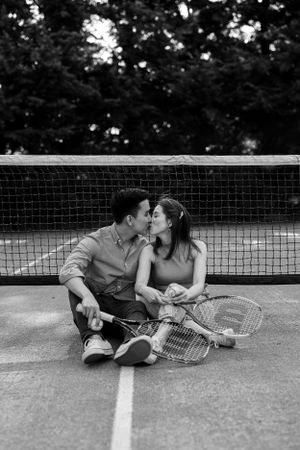 A couple shares a sweet moment while sitting on a tennis court with rackets beside them.