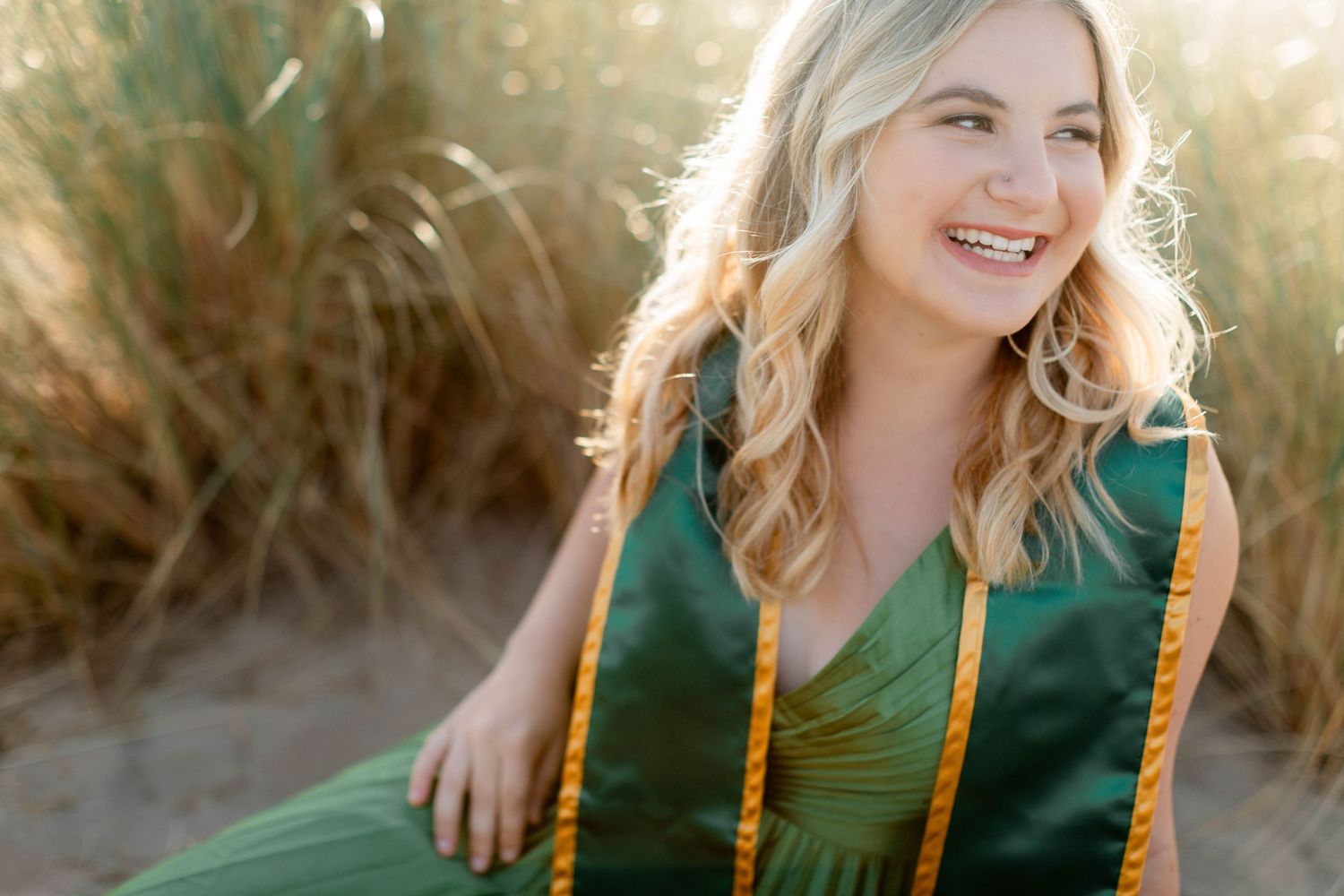 A college senior in a green dress poses outdoors during golden hour with sunlight creating a warm, natural glow.