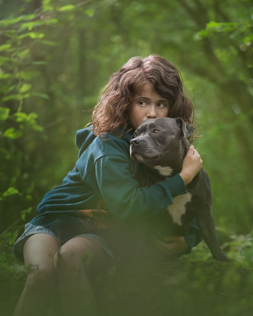 A person in a green jacket hugs their black cat in a lush forest setting.