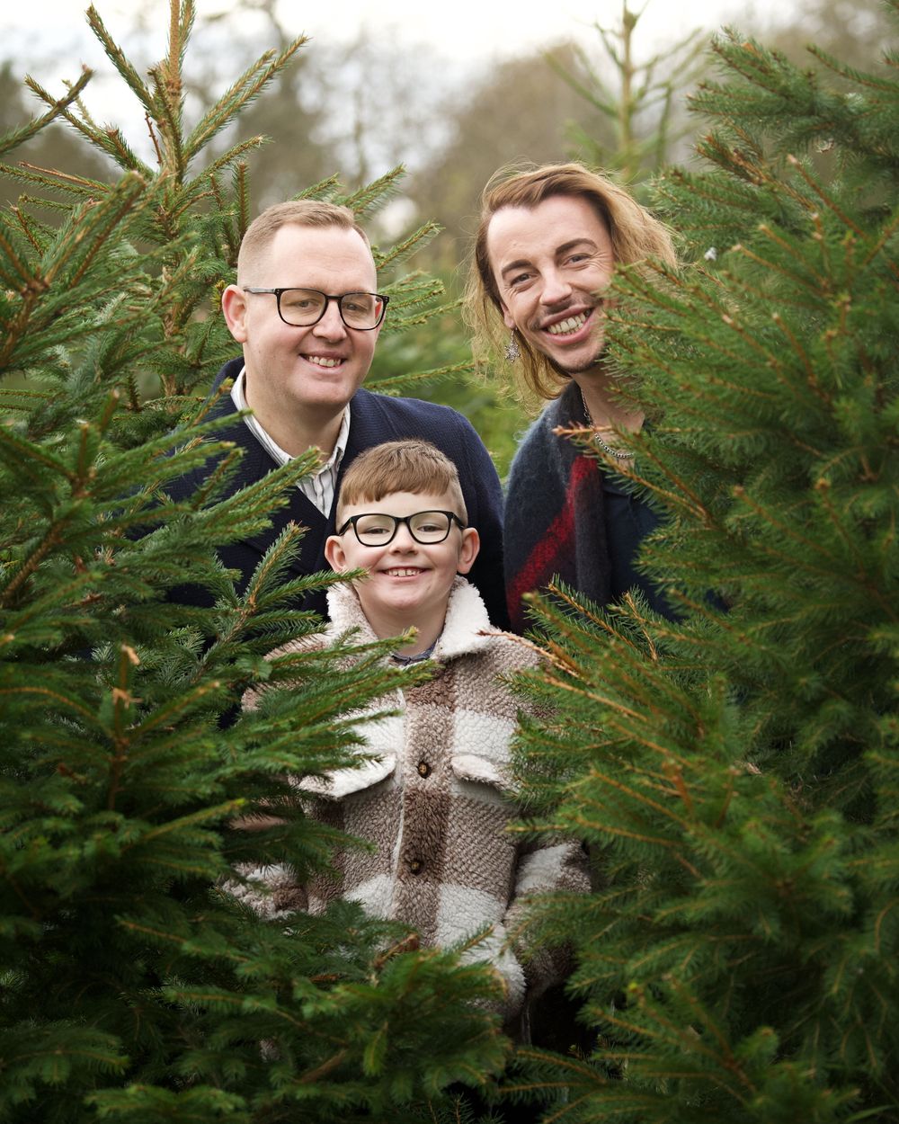 A family poses together between green Christmas trees during the holiday season.