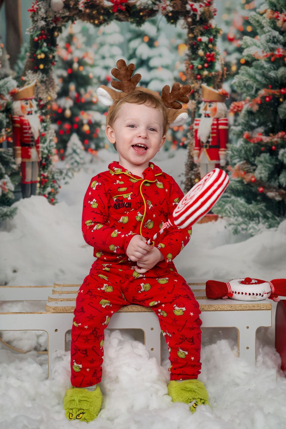 A child in red reindeer pajamas sits on a bench surrounded by Christmas decorations.