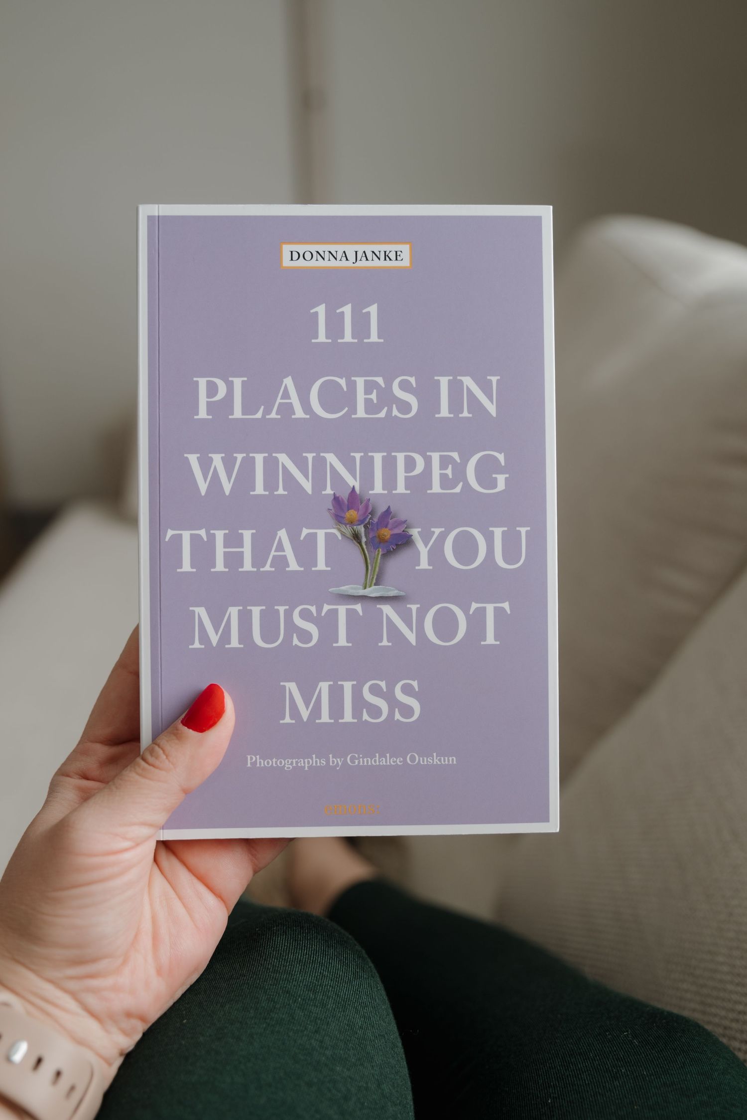 A hand holding a purple book titled '111 Places in Winnipeg That You Must Not Miss' against a soft neutral background.