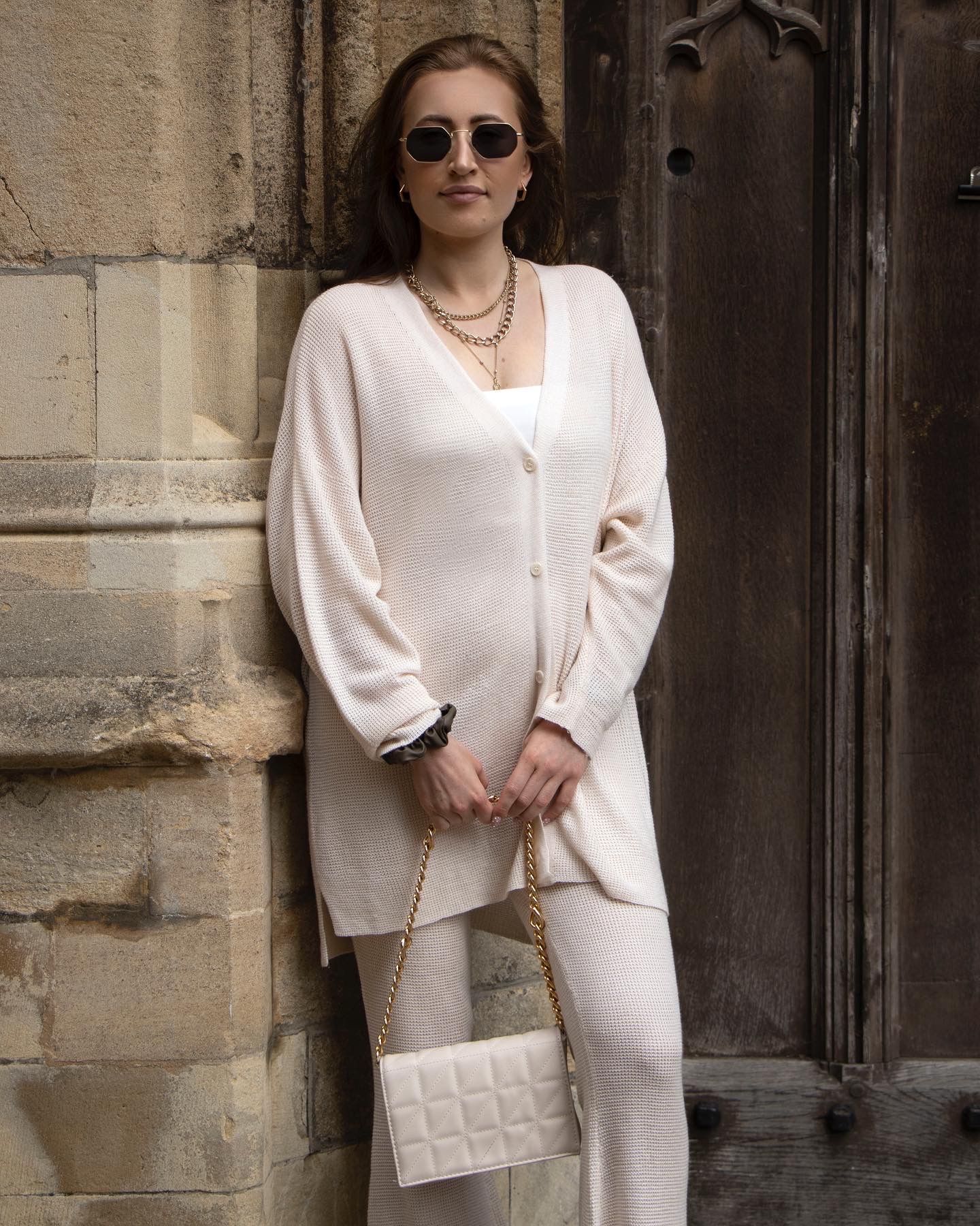 Fashionable woman in cream colored outfit poses against stone wall wearing sunglasses and holding quilted handbag.