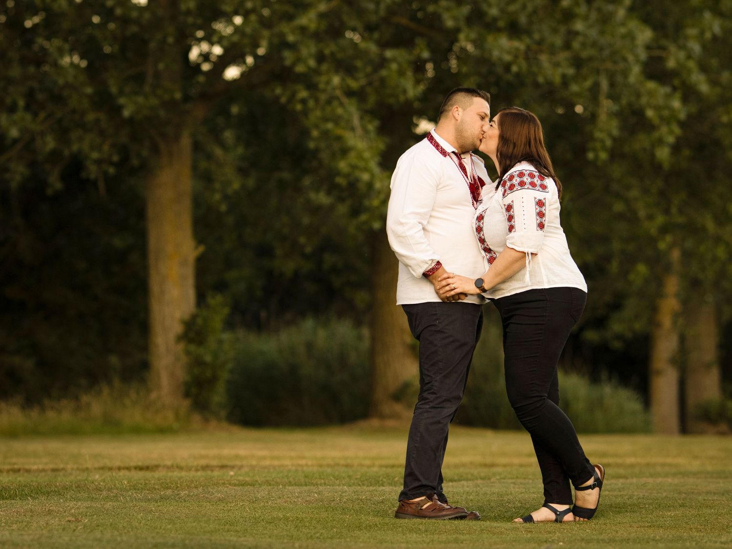 A romantic couple wearing matching white tops embrace on a grassy field during golden hour.