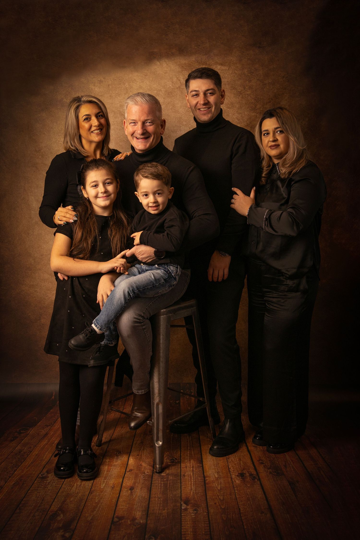 Extended family group photo with warm lighting on wooden floor backdrop.
