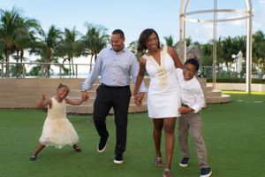Joyful family of four walking and holding hands on a green lawn with tropical palm trees behind them.