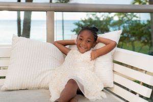 Adorable young girl in a white dress relaxing on a porch bench with pillows, smiling happily.