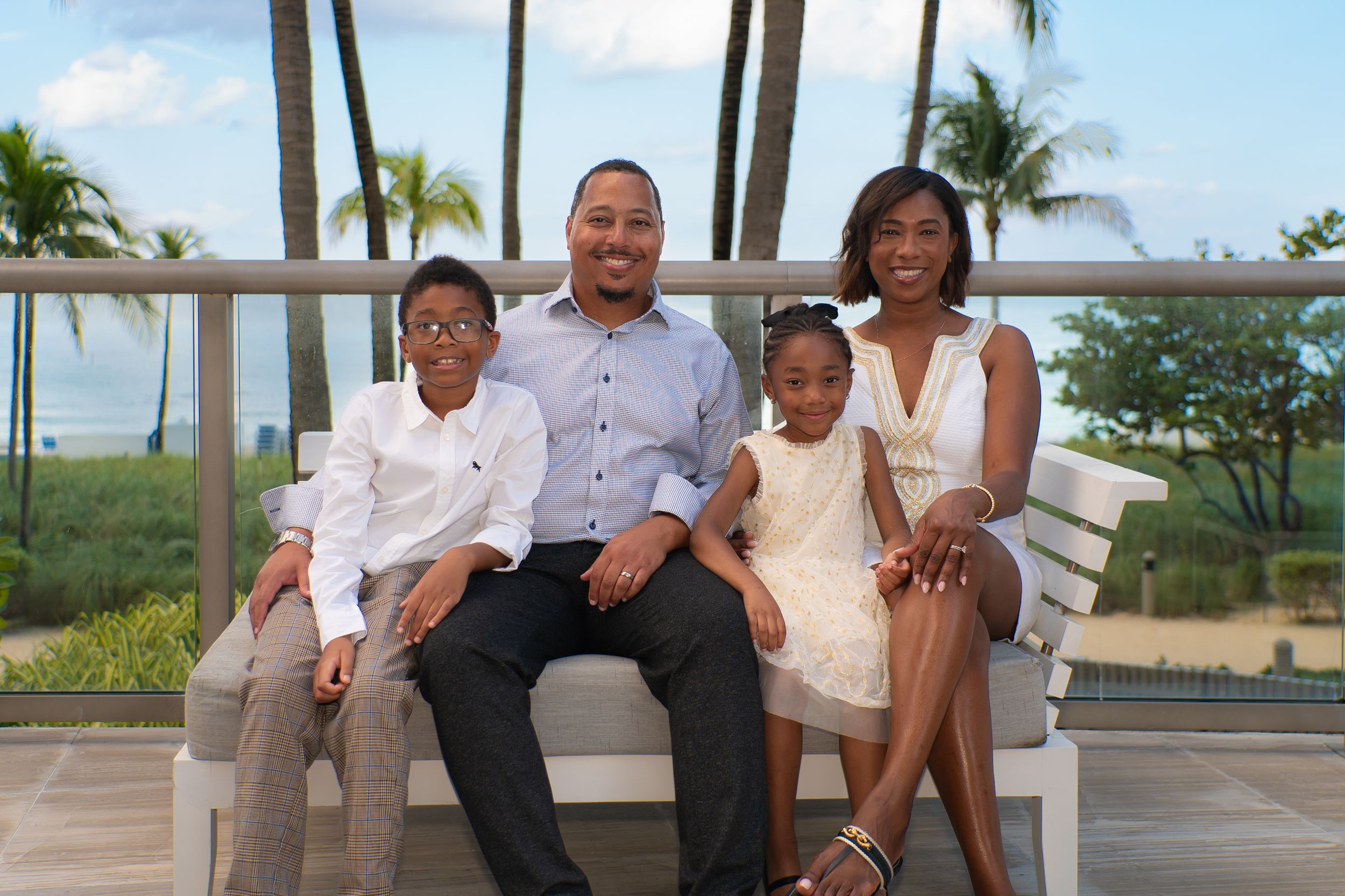 Happy family of four posing together on a bench with palm trees and ocean view in background.
