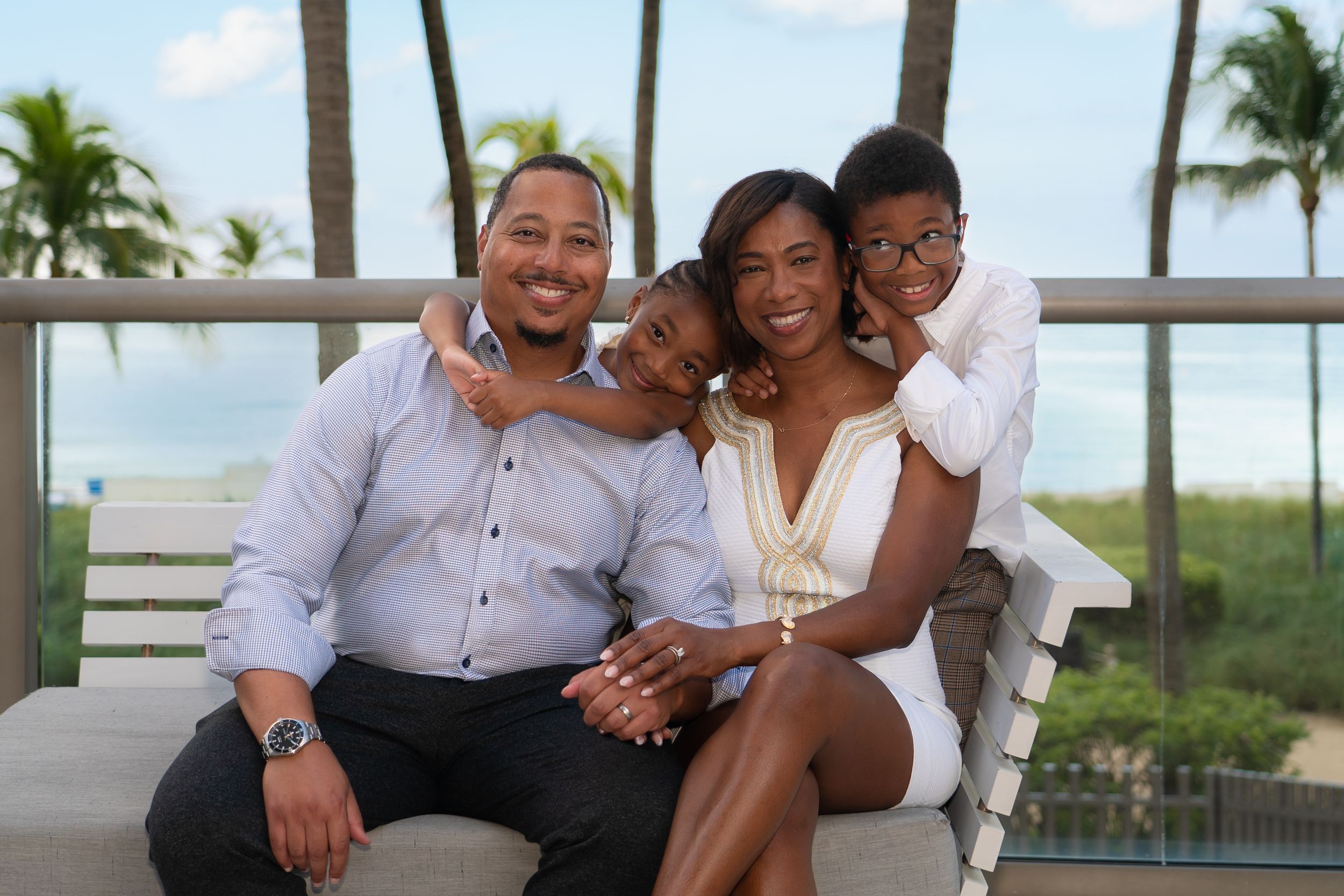 Smiling family of four sitting close together on a bench with a beautiful tropical ocean backdrop.