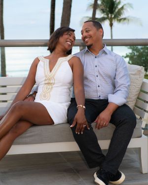 Couple sitting on a white bench, smiling at each other with palm trees and ocean in the background.