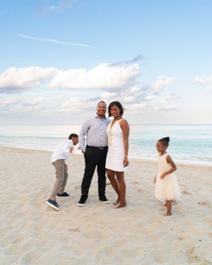 Family of four posing together on a sunny beach with blue sky and ocean backdrop.