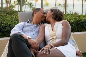 Romantic couple sharing a kiss while seated together on a chair with tropical palm trees in background.