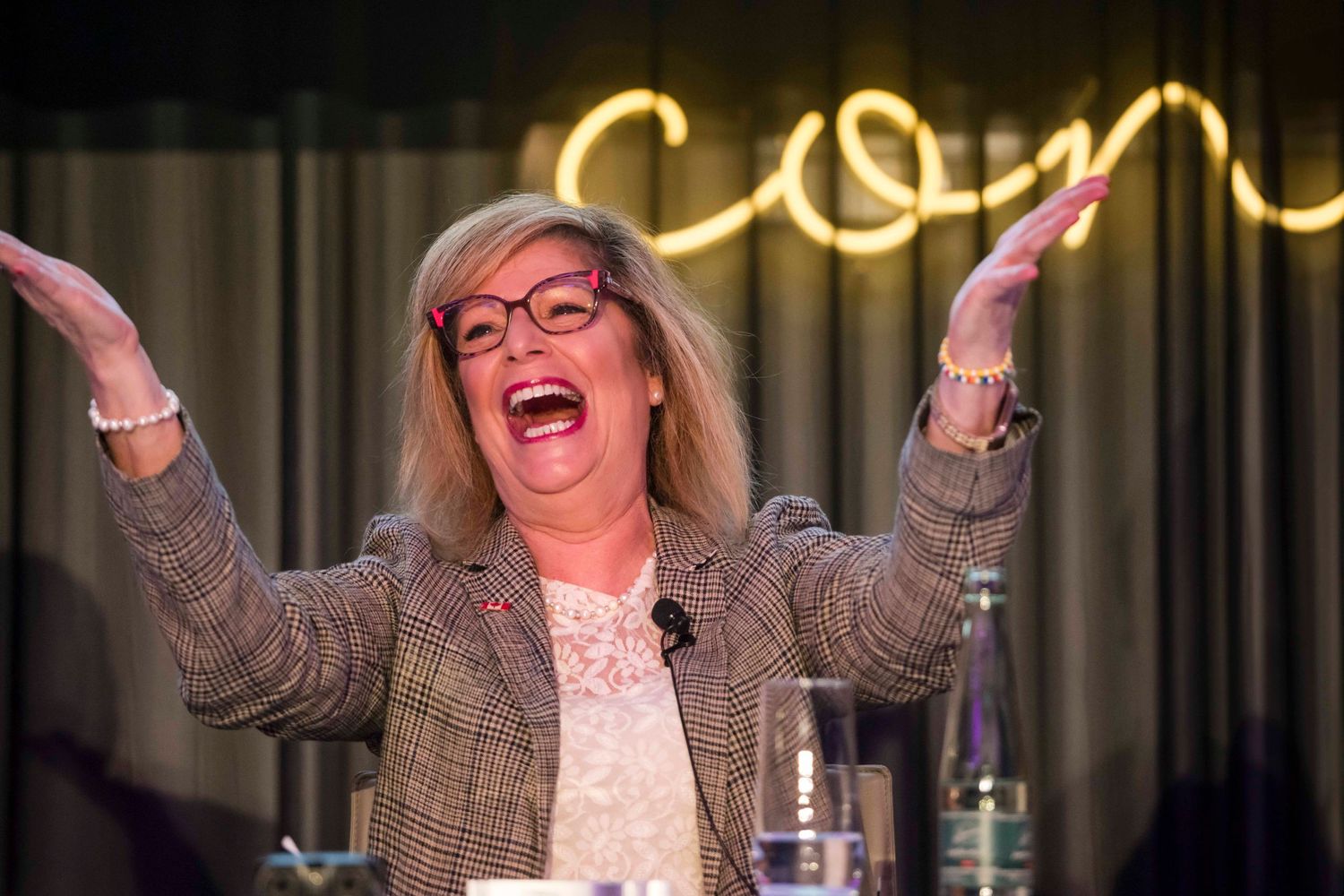 Lively woman in tweed jacket gestures enthusiastically under neon lighting at comedy club.