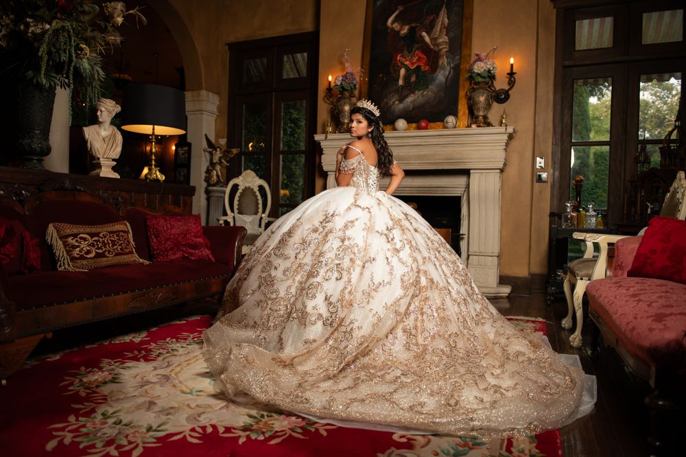 An elaborate white and gold quinceañera dress displayed in a luxurious living room with red velvet furniture.