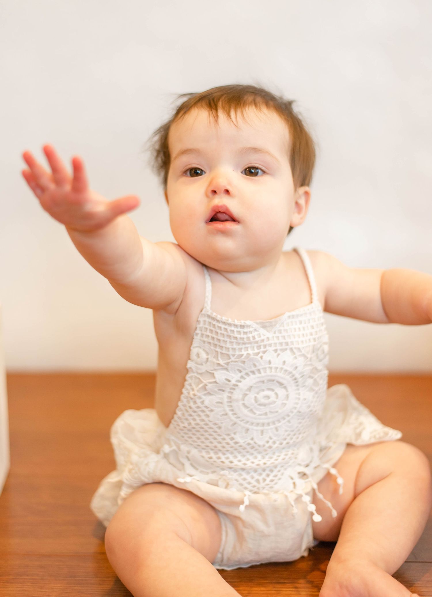 A baby in a white lace romper reaches up while sitting on hardwood flooring.