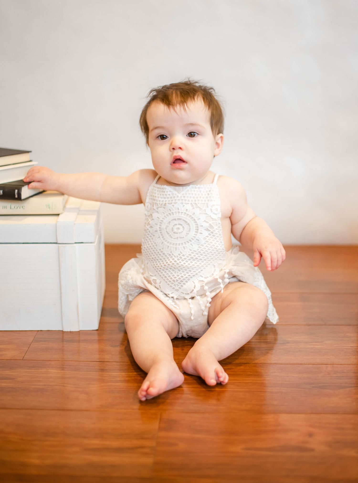 Baby wearing white lacy outfit sits on wooden floor next to white storage boxes.