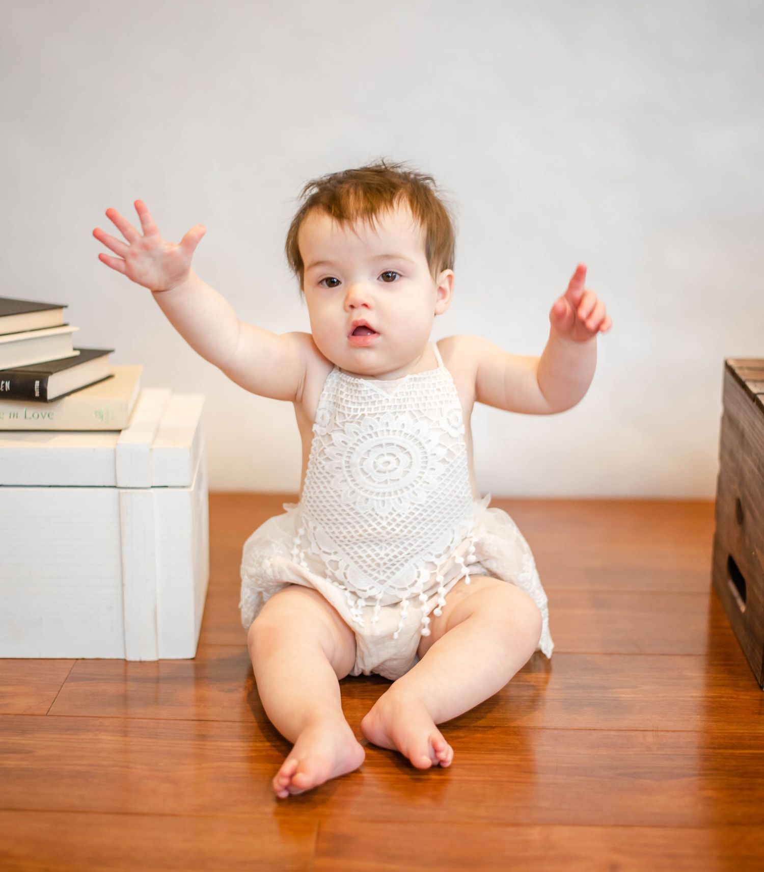 Baby in white lace outfit raises arms while sitting on hardwood floor near books.