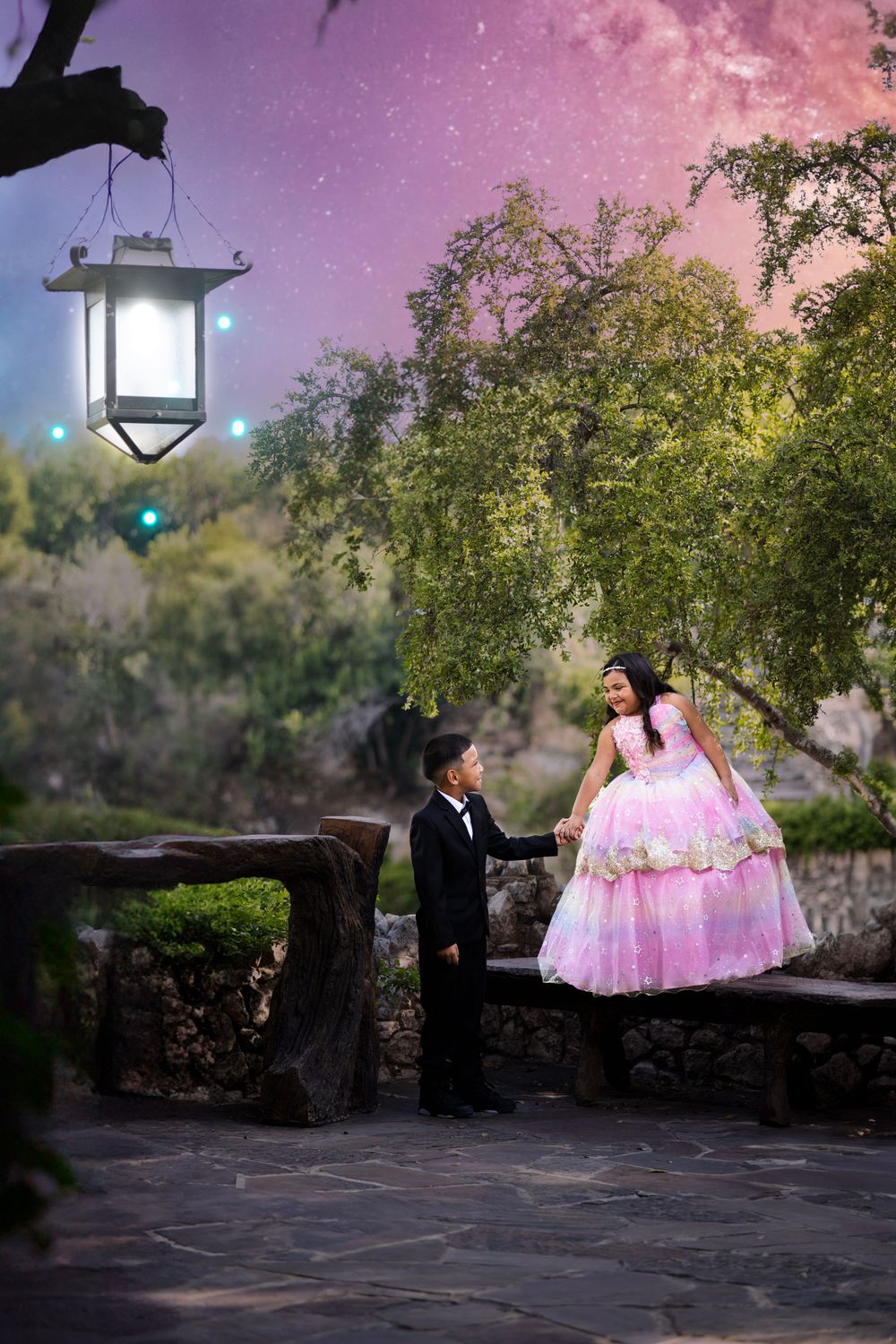 Two figures in formal attire standing by a stone wall at dusk with glowing lantern and pink sky.