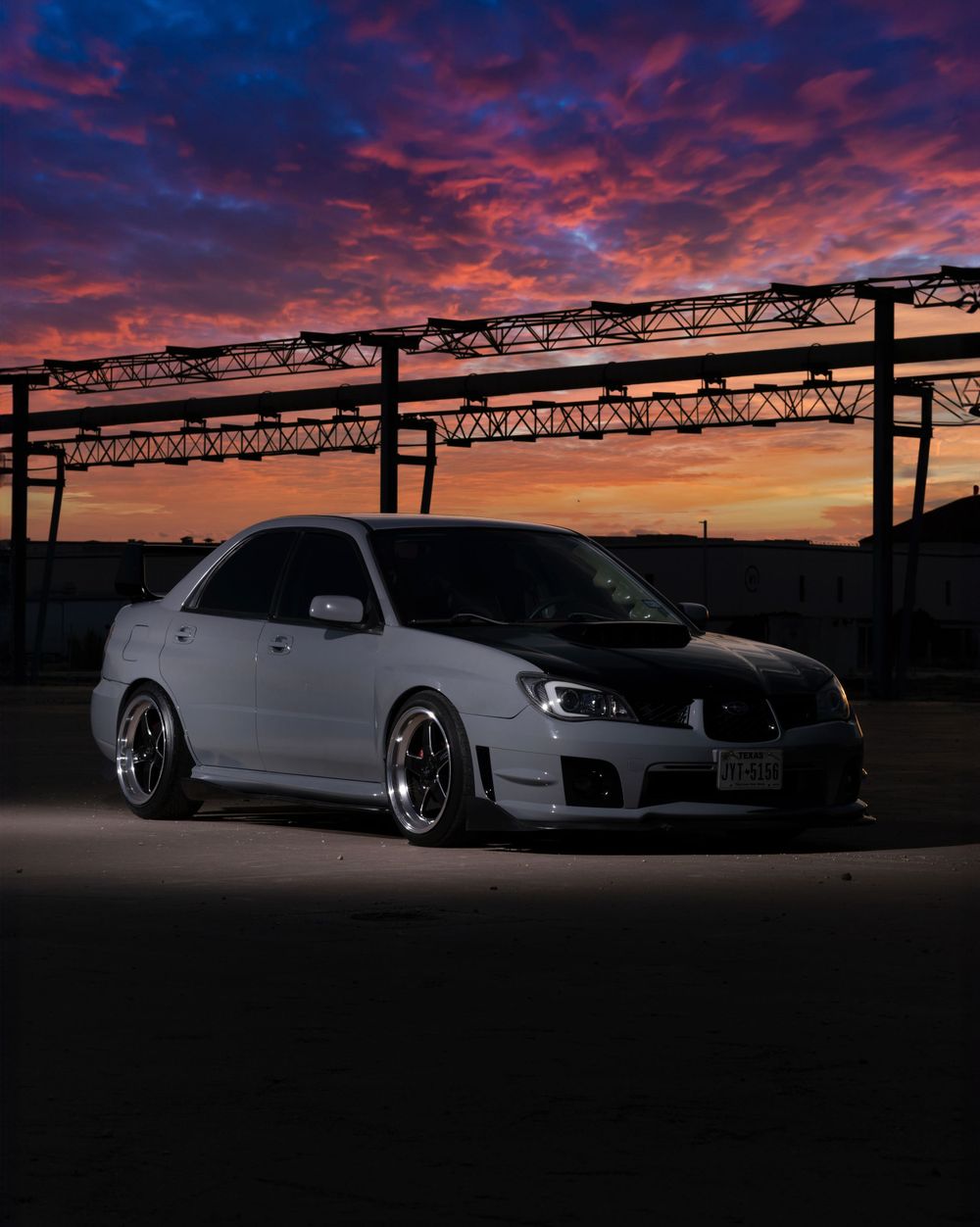 Modified white Subaru WRX STI parked under industrial beams during dramatic sunset.
