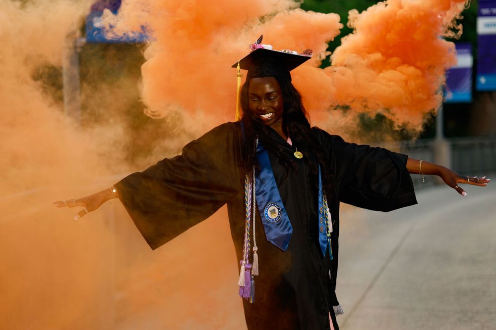 Graduate in cap and gown poses with orange smoke effects in urban setting.
