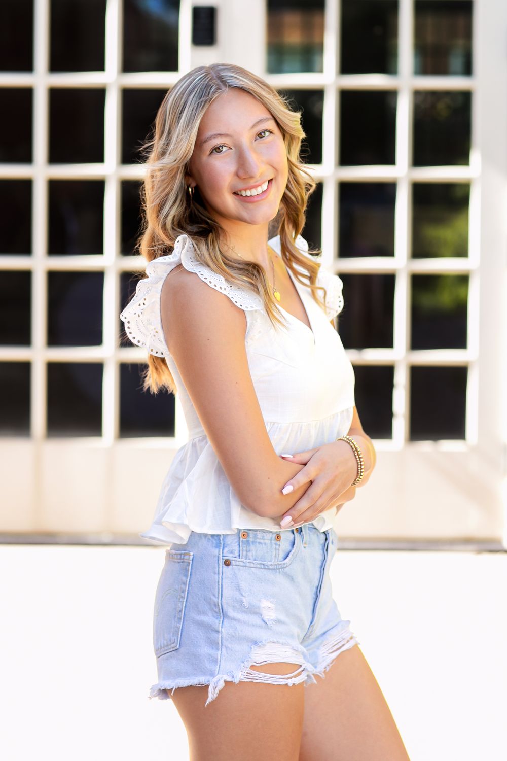 Young woman in white top and denim shorts poses casually against white building exterior.