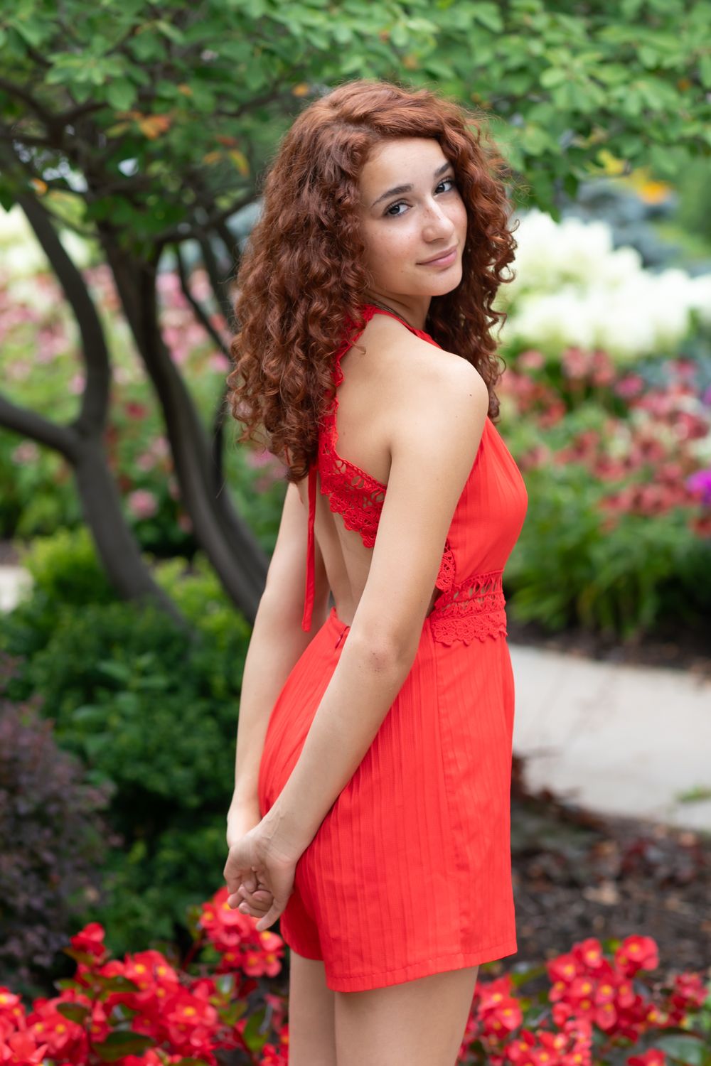 Woman in a bright red sleeveless top poses in a garden with blooming red flowers.