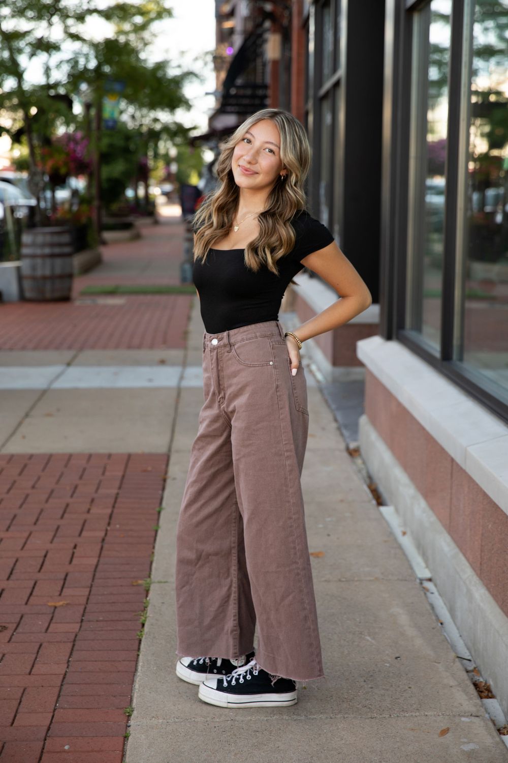 Person wearing brown pants and black sneakers stands on brick sidewalk near storefronts.
