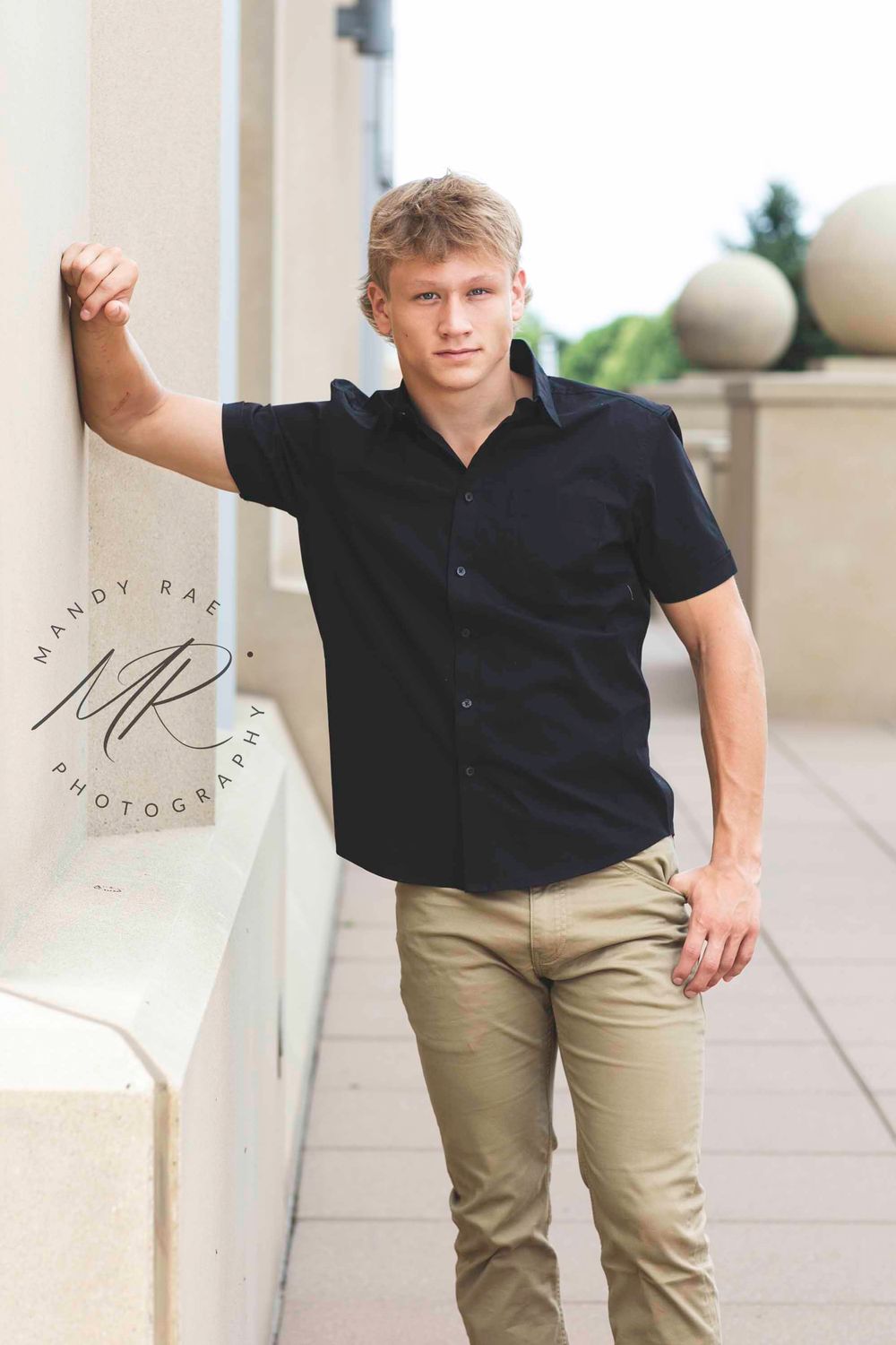 Young person in casual black polo and khaki pants poses against light colored columns outdoors.