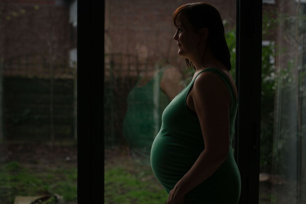 A silhouetted expectant figure stands near a window in dim lighting during pregnancy.