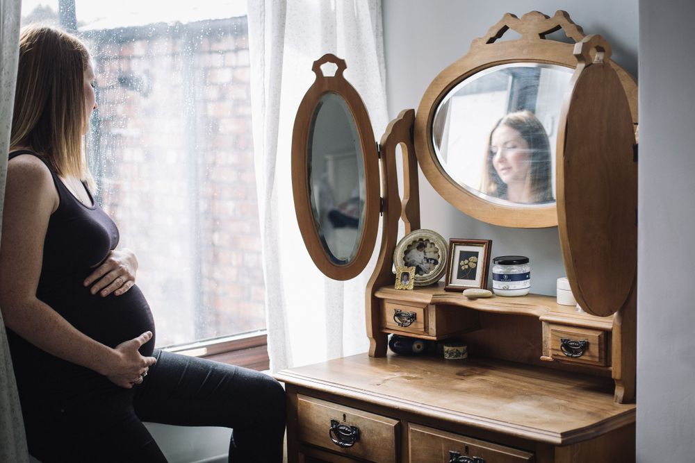 A pregnant silhouette stands before a vintage vanity mirror in a softly lit room.