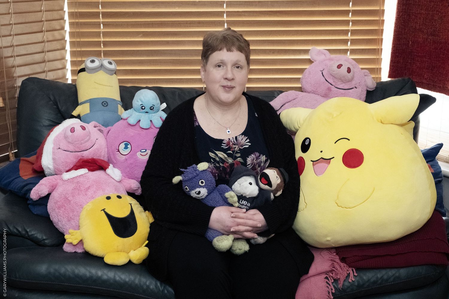 Someone on a dark couch surrounded by colorful plush toys including Pikachu, Minions and pink characters.