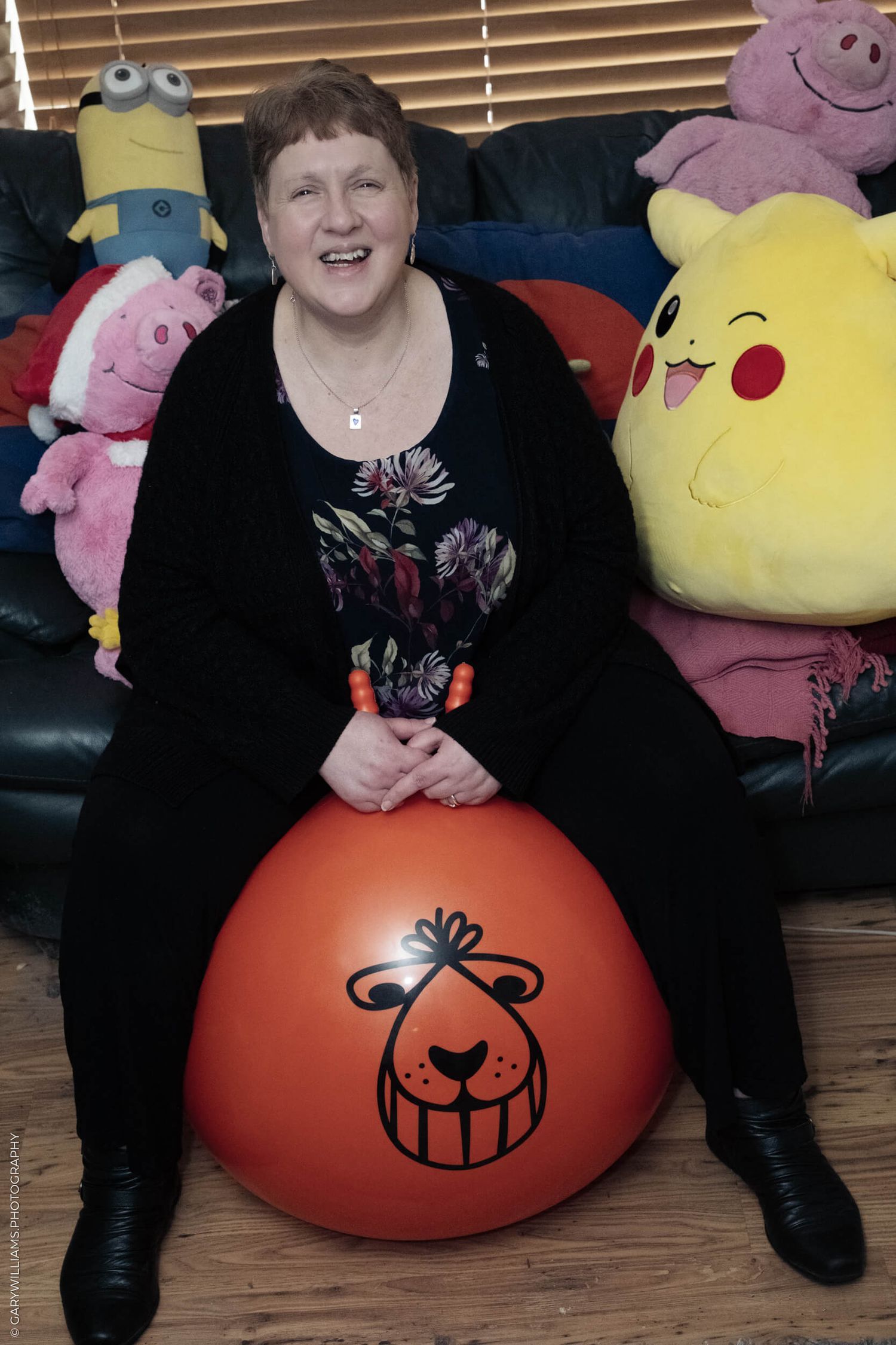Someone sitting on a couch with stuffed toys including Minion, Pikachu, and pink plush animals and a red bounce ball.