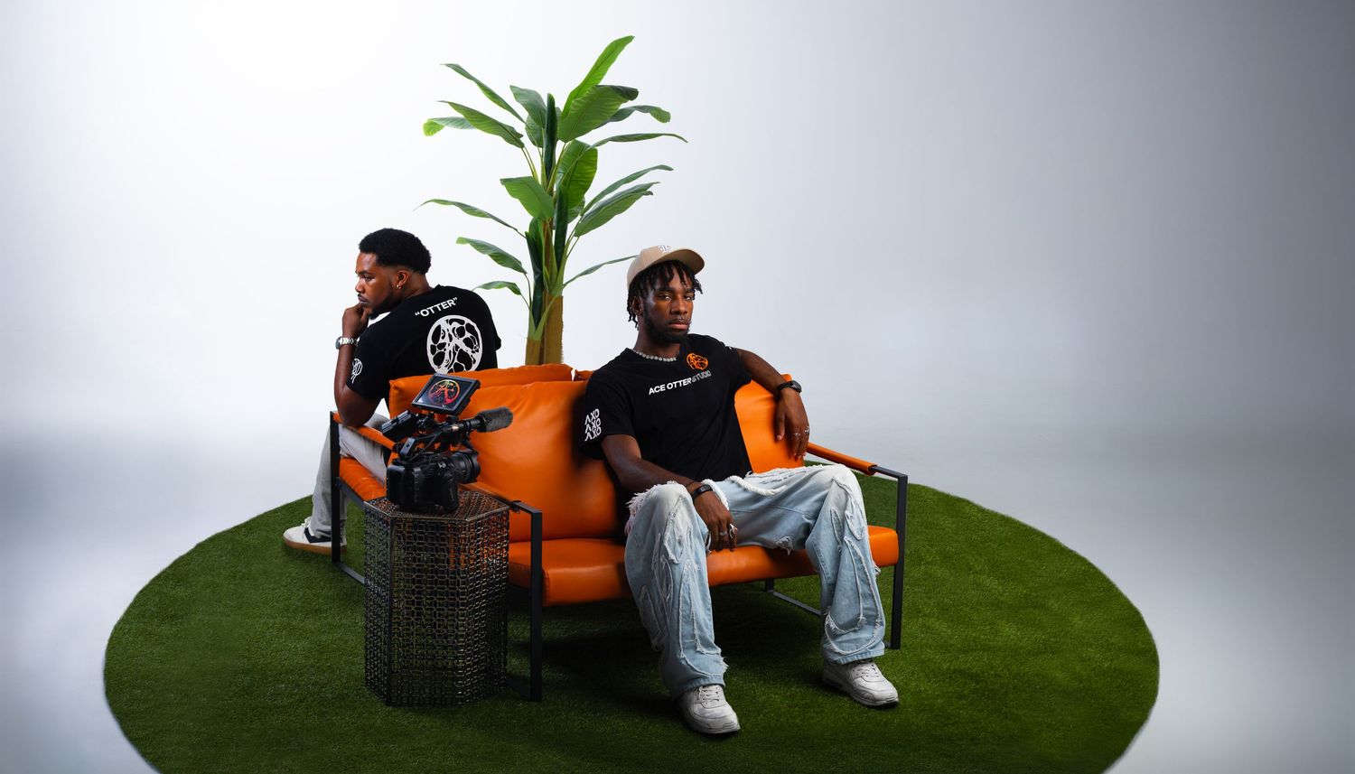 Two men sitting on an orange sofa on artificial grass with a tropical plant, wearing streetwear outfits.