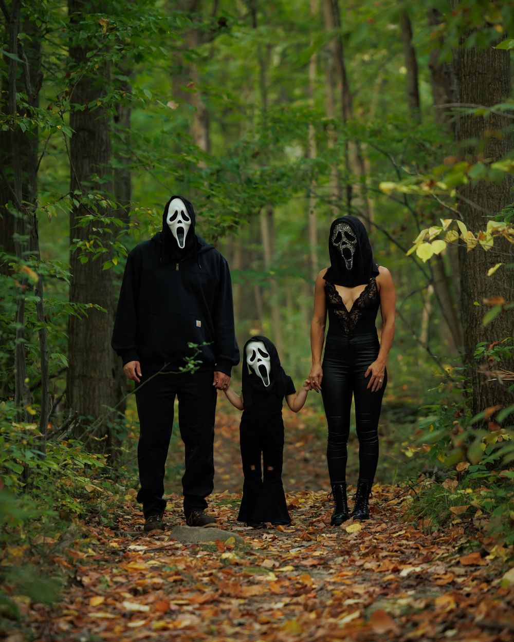 A group wearing black hooded costumes and ghost face masks walks through a misty autumn forest path covered in fallen leaves.