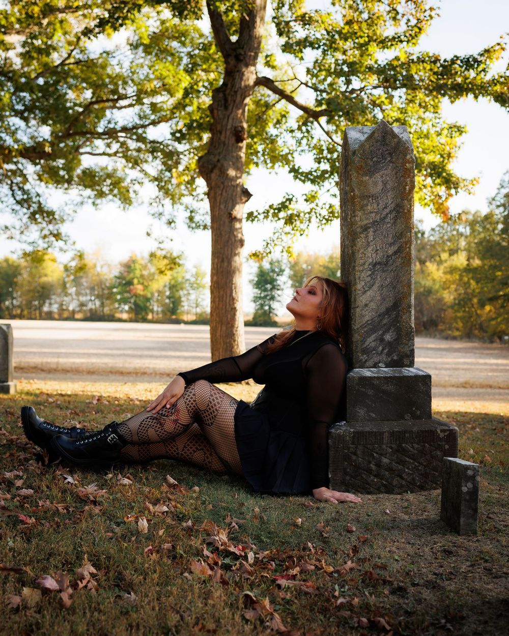 A figure reclines against a tree trunk in autumn, surrounded by fallen leaves and dappled sunlight in a peaceful setting.