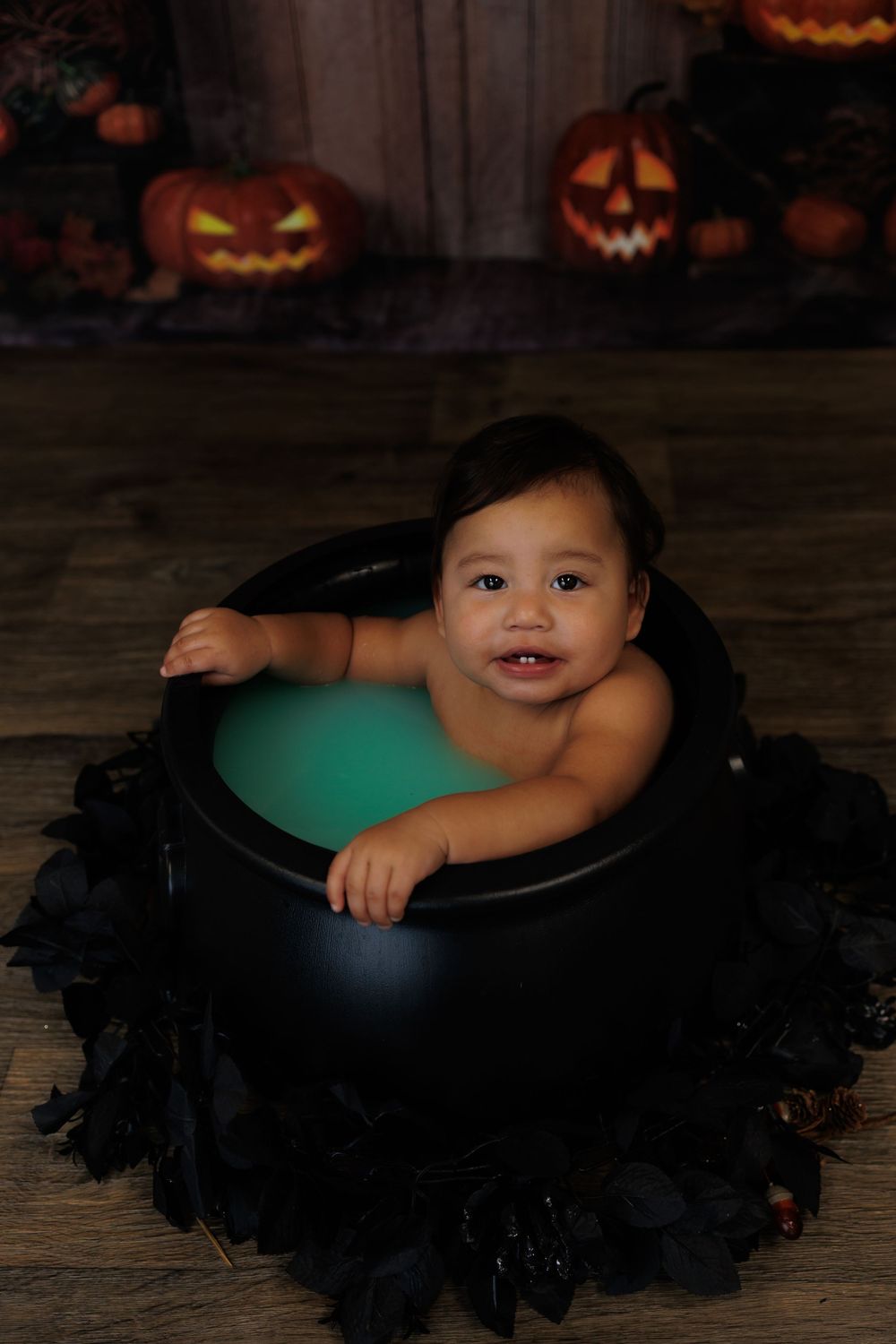 A child wearing a black tutu dress sits against a dark Halloween backdrop with glowing jack-o'-lanterns.