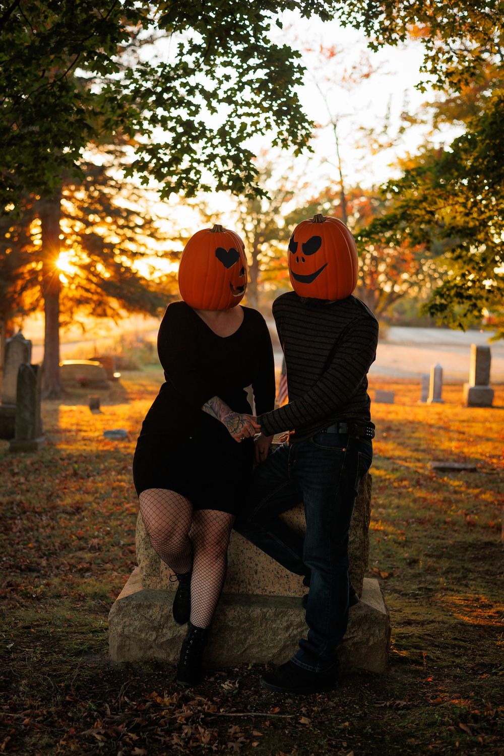 Two people with pumpkin heads sit together on a bench at sunset in an autumn cemetery scene.