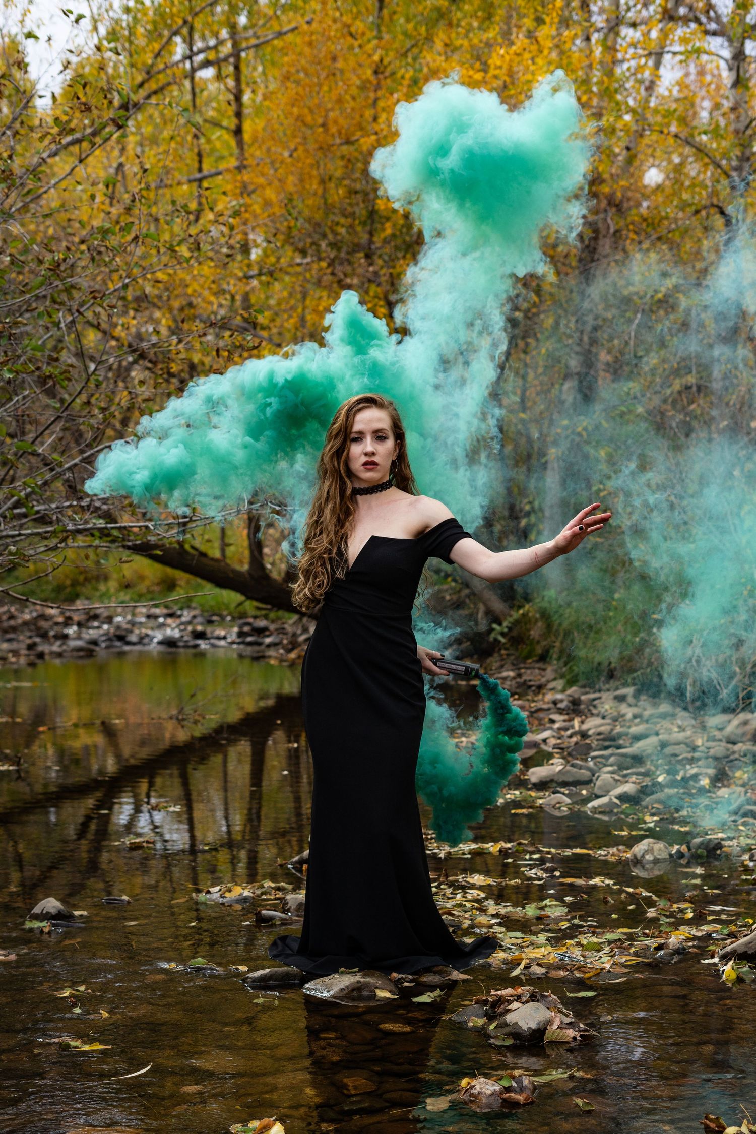 A person in a black dress stands in a stream surrounded by yellow autumn trees and plume of dramatic teal smoke.