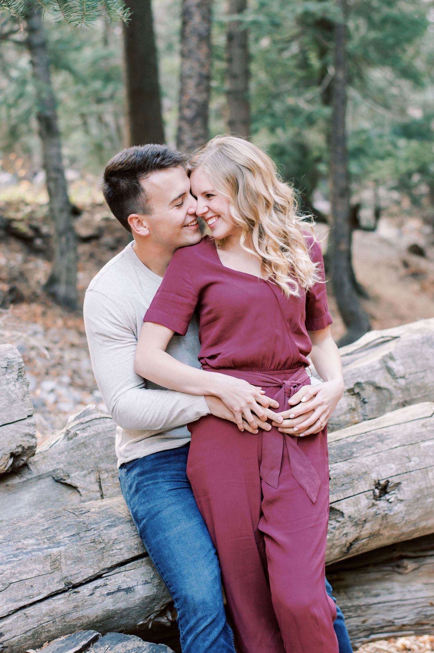Romantic outdoor forest engagement photo of couple sitting on fallen log in natural wooded setting.