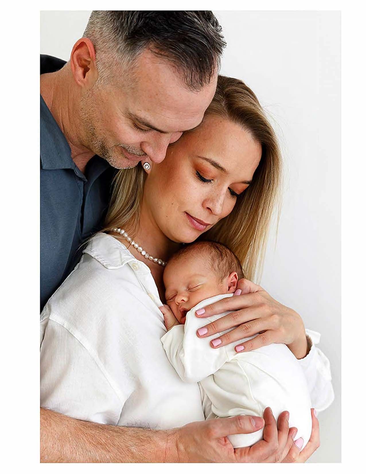 A tender intimate family moment with two adults embracing a sleeping newborn baby in white clothing.