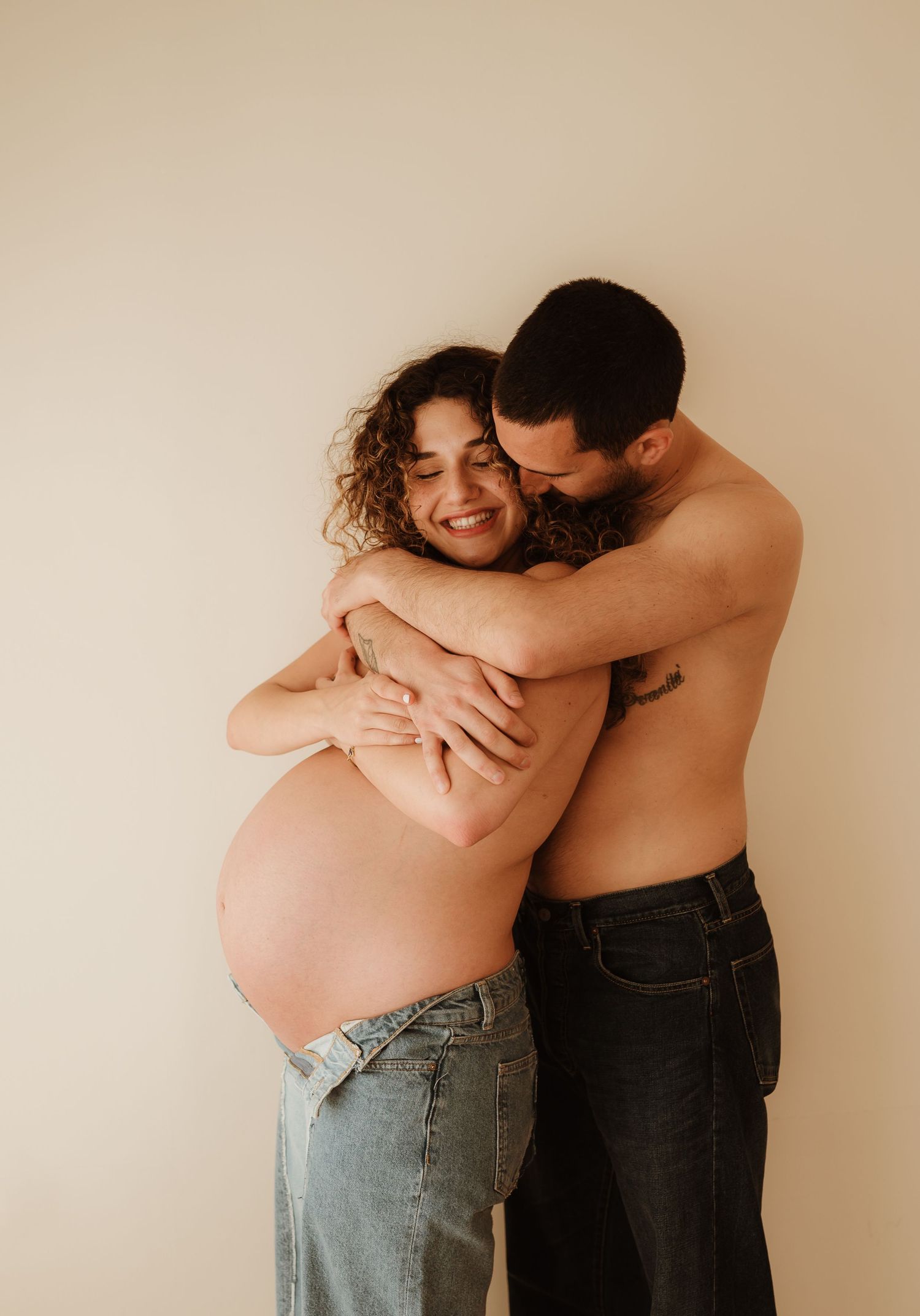 A shirtless couple embraces in a warm emotional maternity photo against a beige wall.
