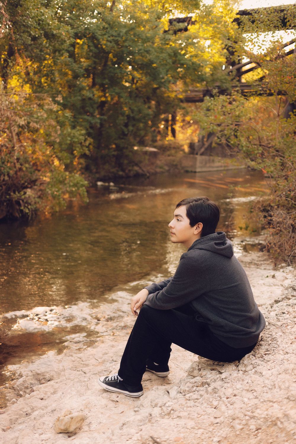 Someone wearing a dark hoodie sits contemplatively by a peaceful stream in autumn with golden foliage overhead.