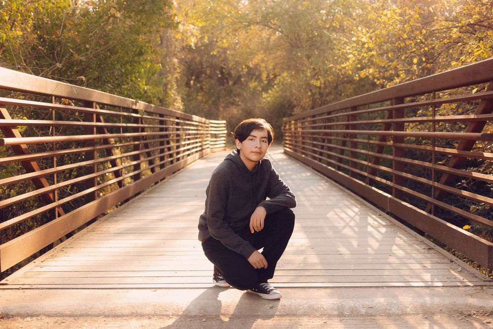 A figure crouches on a wooden bridge walkway surrounded by autumn foliage in golden afternoon light.