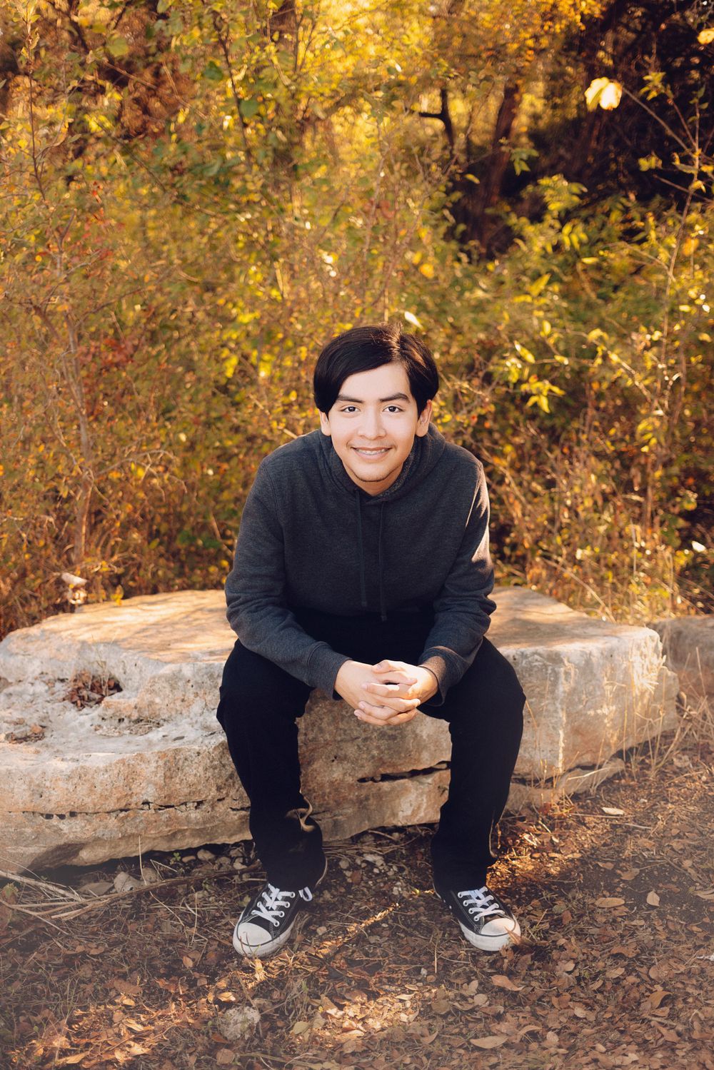 A person in a black sweater and pants sits on a large rock amidst autumn foliage in a park or forest setting.