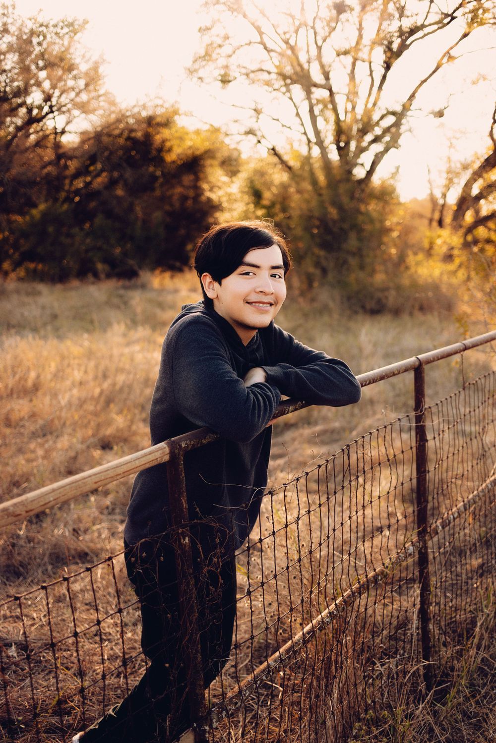 A person in black clothing leans against a rustic wire fence at sunset in an autumn field with golden light and trees.