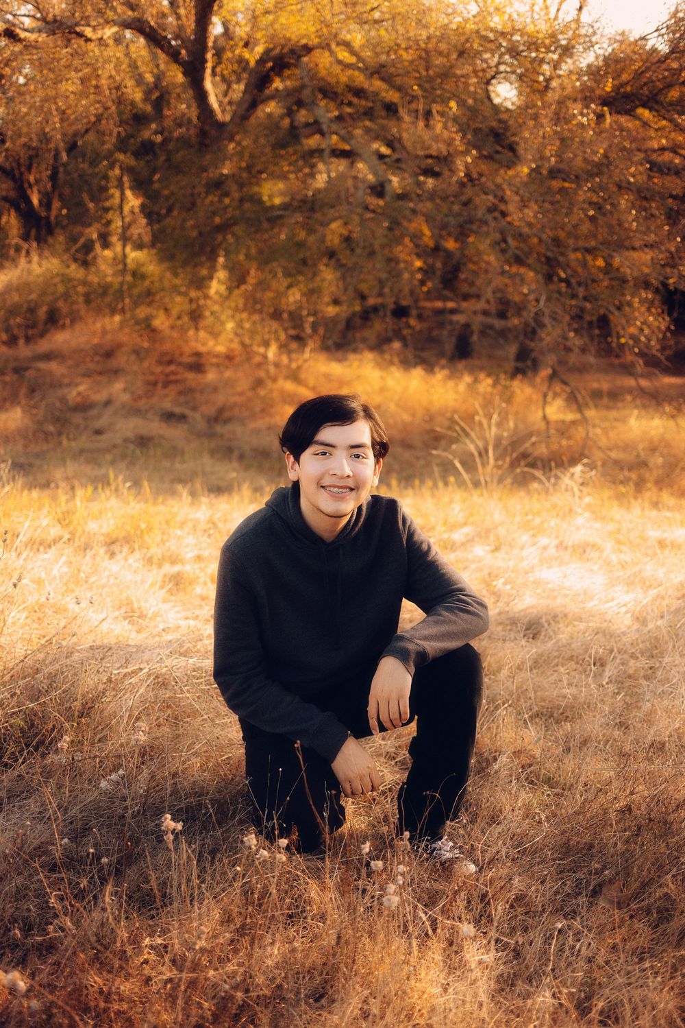 A person in a black sweater kneels in golden autumn grass with sunlit trees in the background.