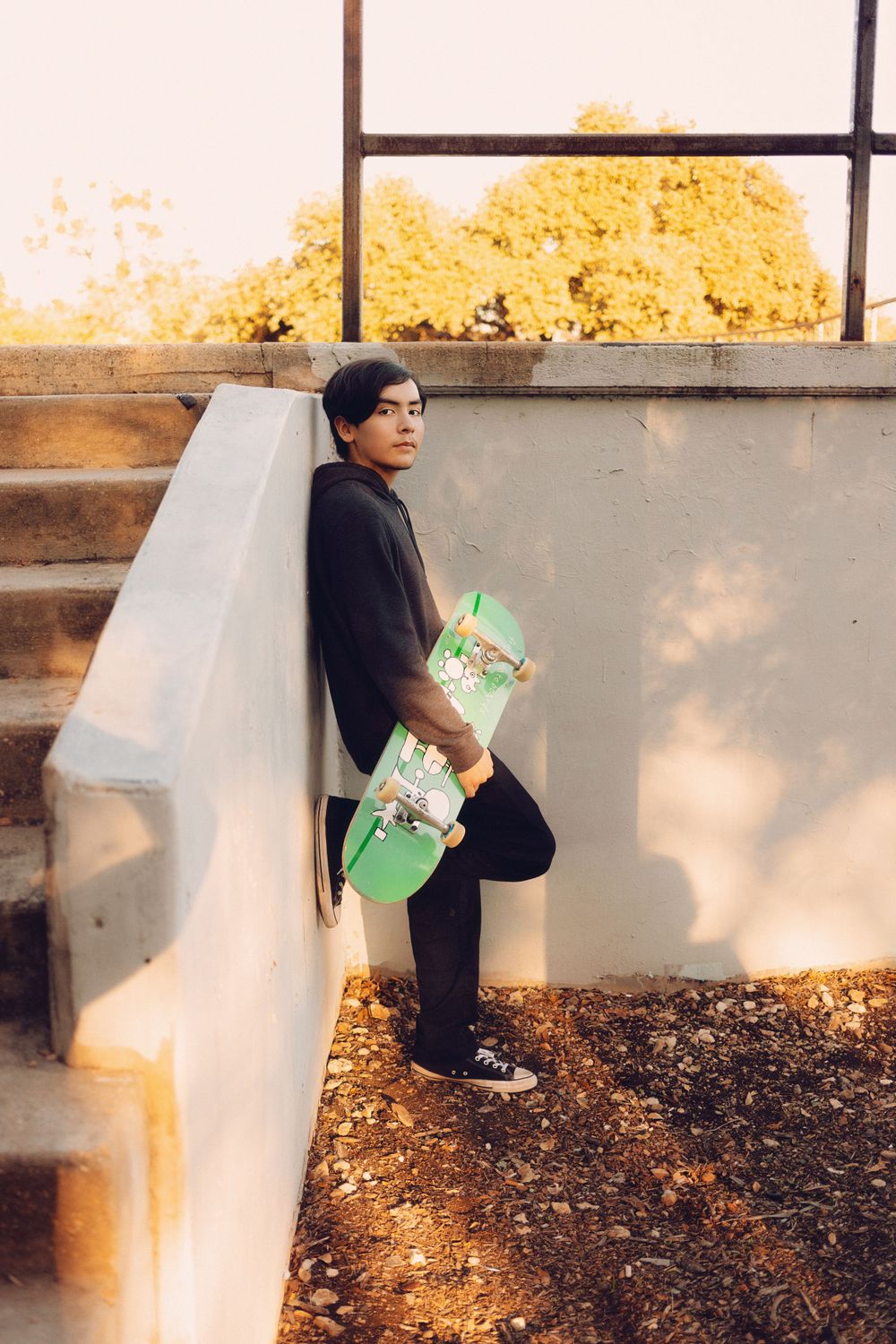 A skateboarder in a black hoodie leans against a concrete wall holding a mint green skateboard during golden hour.