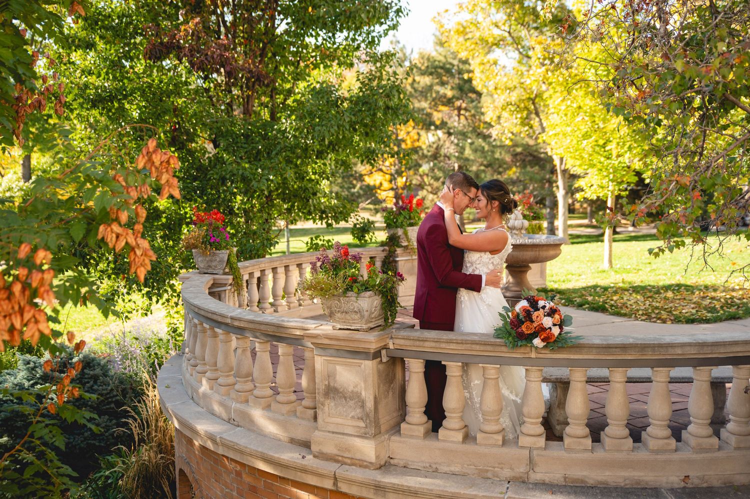 A couple shares a romantic moment on an ornate stone balustrade surrounded by autumn foliage in a garden setting.